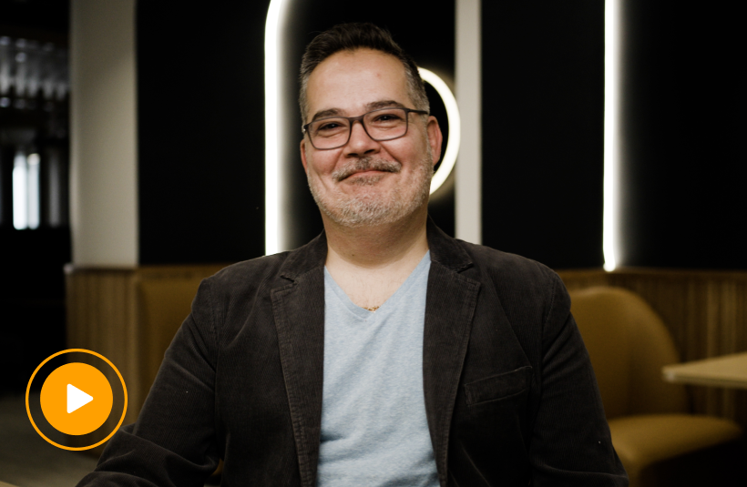 Fabio Victorino smiles at the camera, wearing glasses and a dark blazer over a gray t-shirt, seated in front of glowing vertical light panels. An orange play button icon overlays the bottom left, indicating a video.