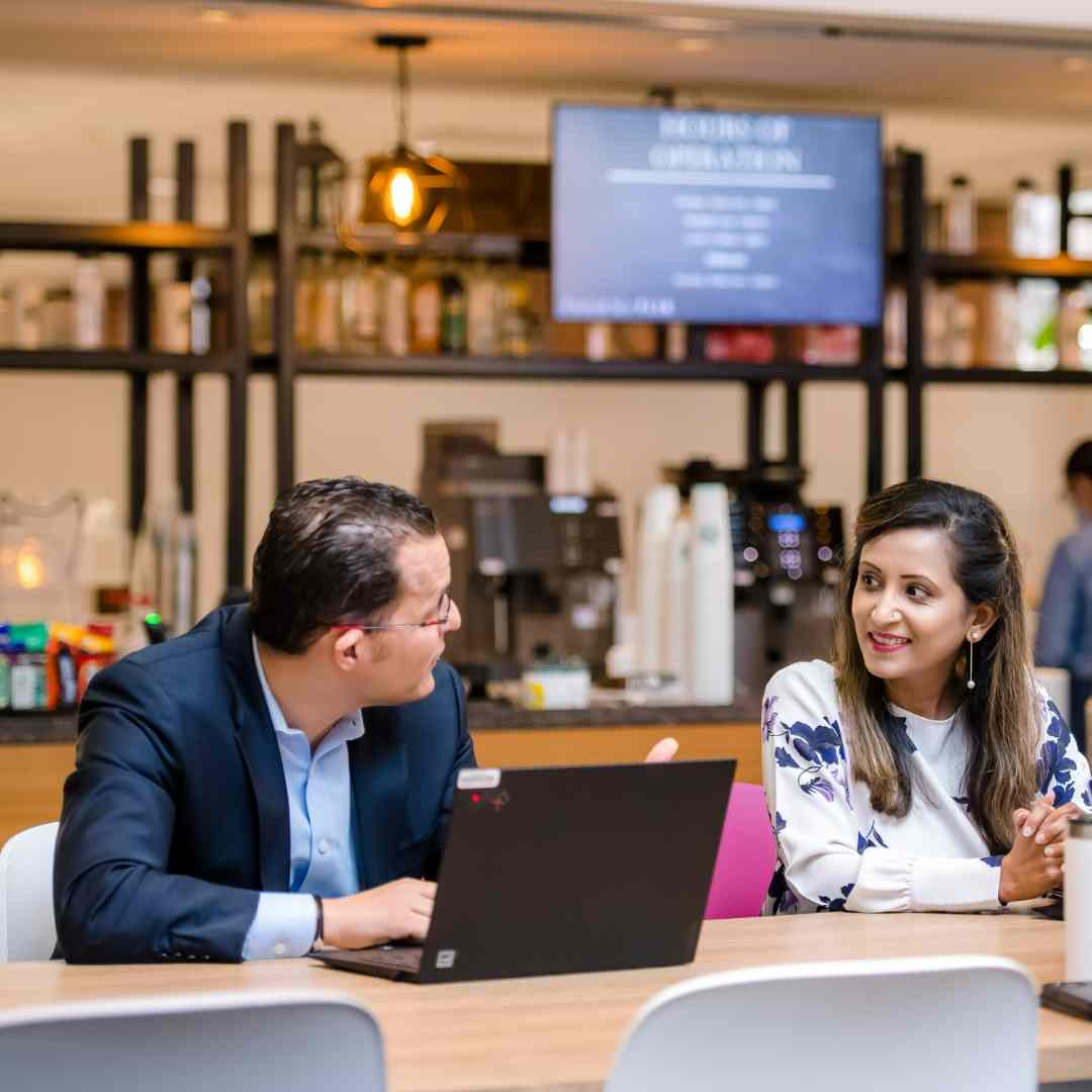 Man using laptop in modern cafe setting talking to female colleague