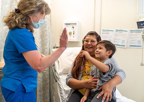 A child sits on the lap of their guardian in an exam room while high fiving a UNMH staff member