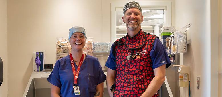 Marjorie, an X-ray Tech, stands to the left of Dr. Wharton, Orthopaedic Trauma Surgeon. They're both smiling in scrubs outside an operating room