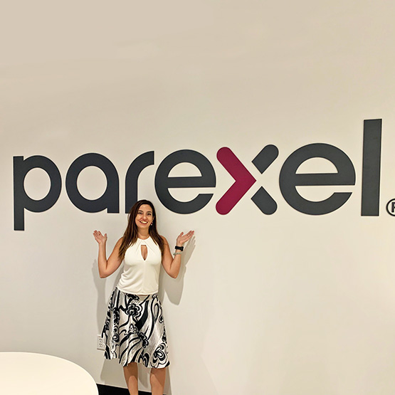 Carolina Andrada, Project Leader, Argentinian, long brown hair, white shirt and black-white skirt, in front of a Parexel sign, pointing with her arms at the sign.