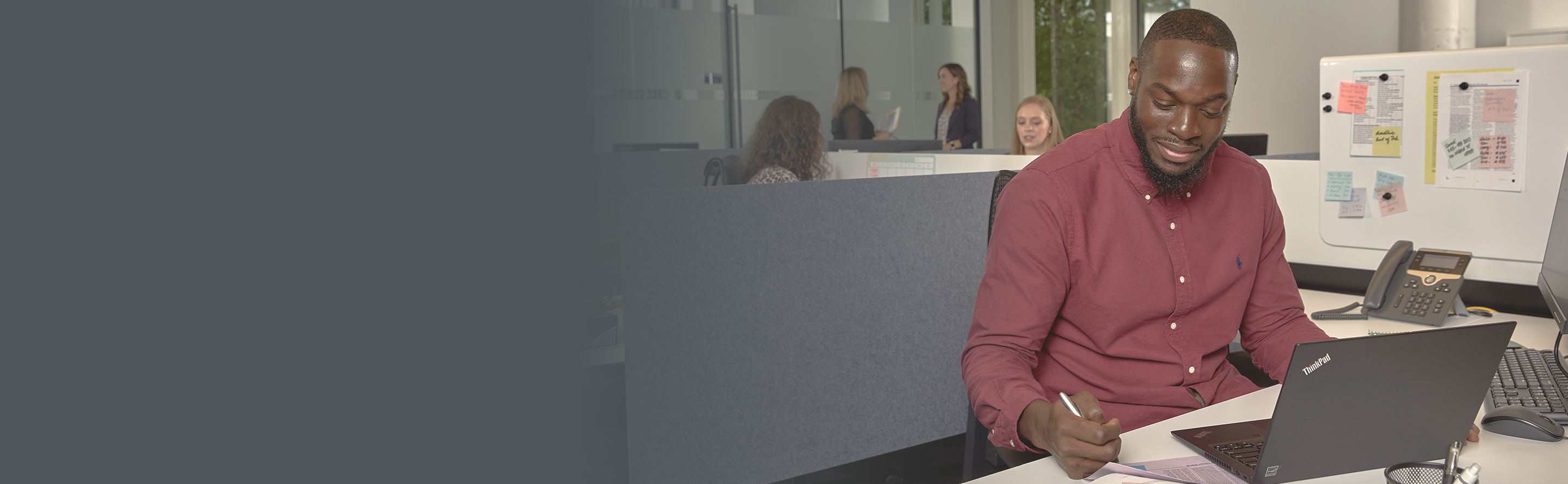 Henry, a Black American Clinical Research Associate, is focused on his work at a desk, utilizing a laptop. He has short hair, a beard, and a red shirt.