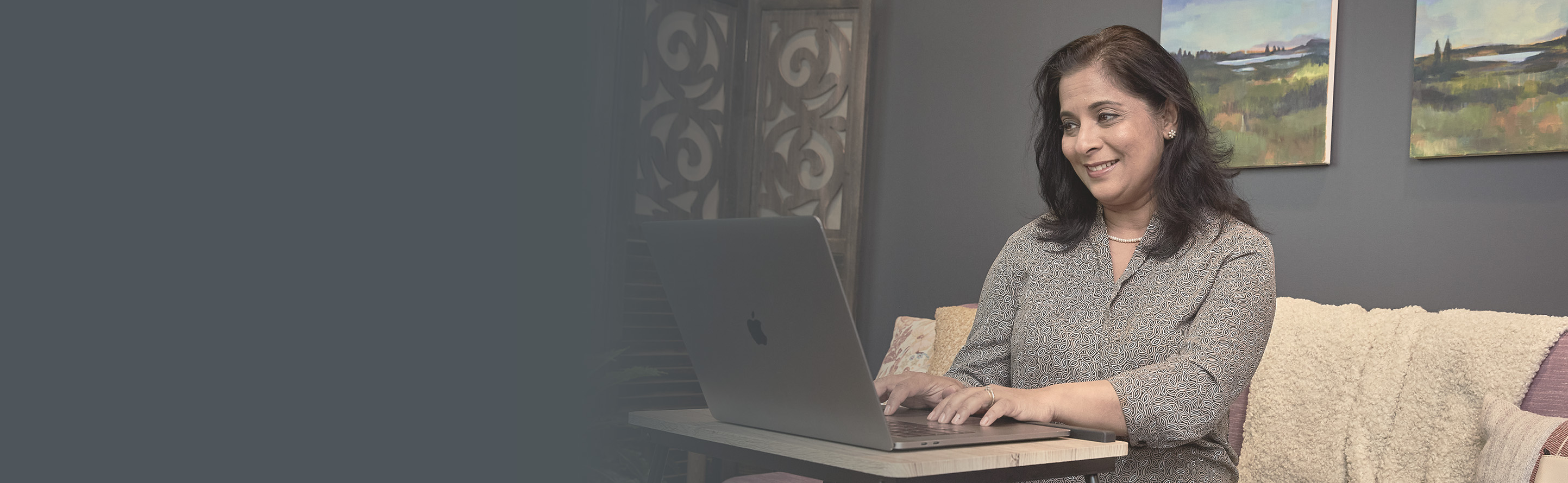 Usree, a Bangladeshi woman and a Statistical Programmer at Parexel, works from home. She sits thoughtfully, using a laptop on a stand. She has long dark hair and wearing a patterned shirt.