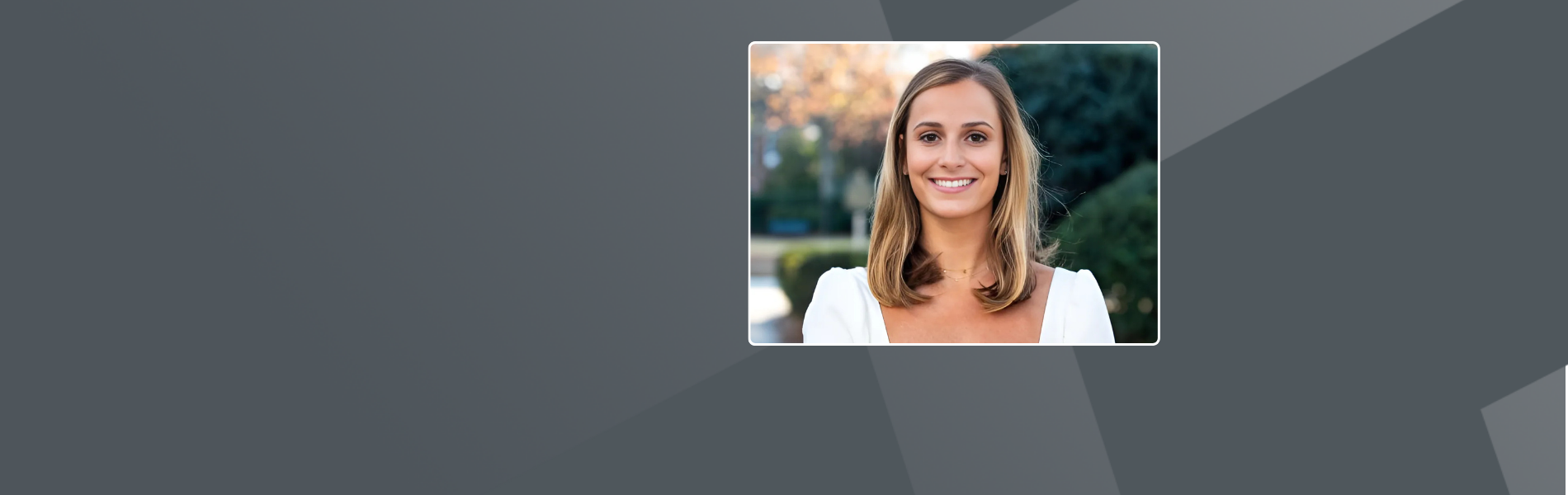 Lillie, a white American, with shoulder-length brown hair and she is wearing a white outfit. It's a portrait picture of her smiling at the camera, against a blurred background of green shrubs and autumn foliage.