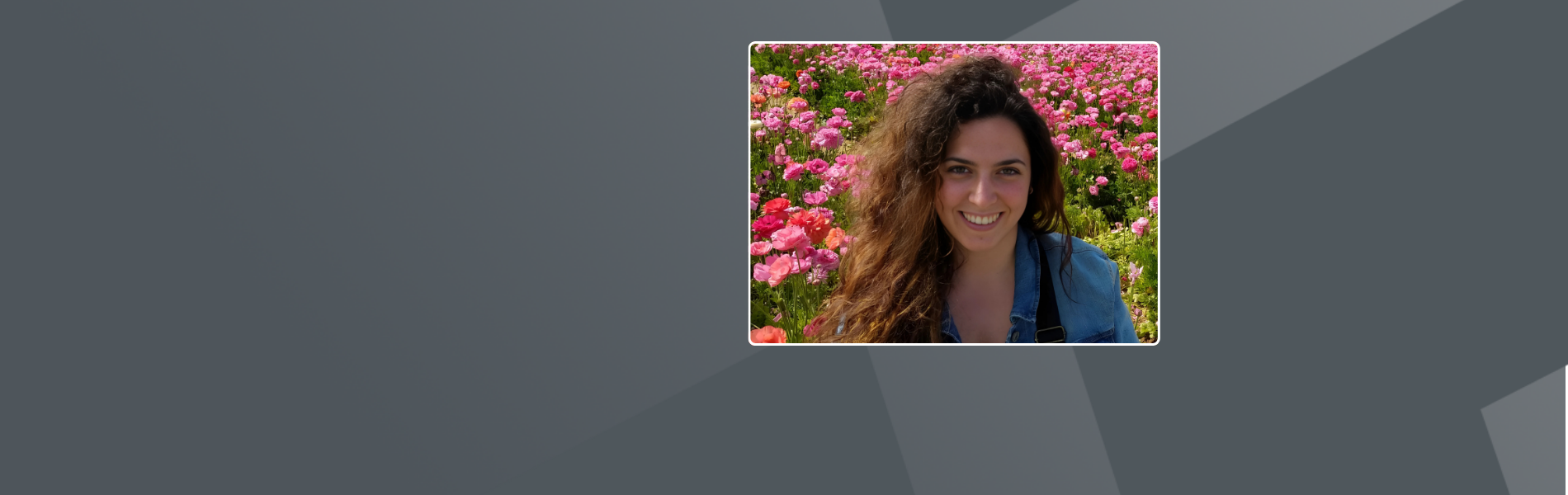Andrea, a Spanish woman, with long curly hair smiles brightly. She is wearing a denim jacket. She's surrounded by a vibrant field of pink and red flowers in full bloom, creating a colorful, cheerful backdrop.