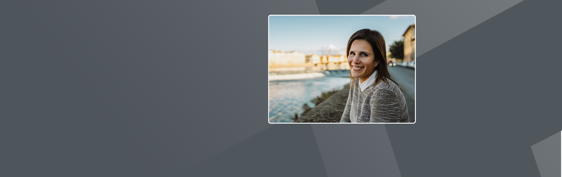 Irene, long brown hair, wearing grey sweater layered over a white collared shirt, sitting on a bench next to the river that flows through Florence, Italy.