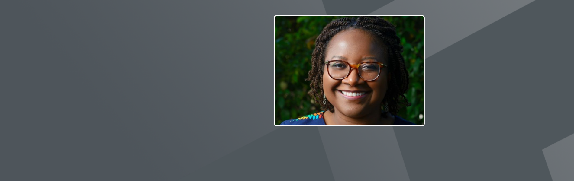 Mwango, a black American woman, with chin long locs, she is waring brown glasses and a patterned shirt, in the background is greenery.