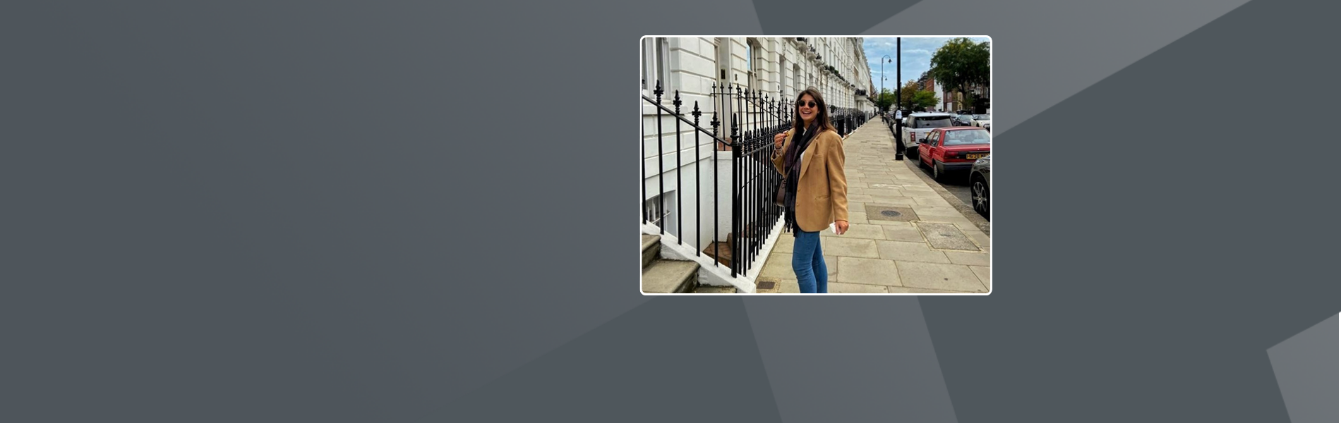 Rebecca, an Italian woman is standing on a London street. She is wearing a beige jacket, a dark scarf and sunglasses. She is standing sideways and is smiling at the camera.