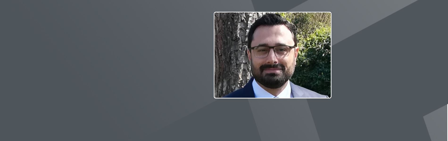 Sinan, a man with brown hair, eyes, and a beard, wearing glasses; while wearing a black suit with a white shirt.
