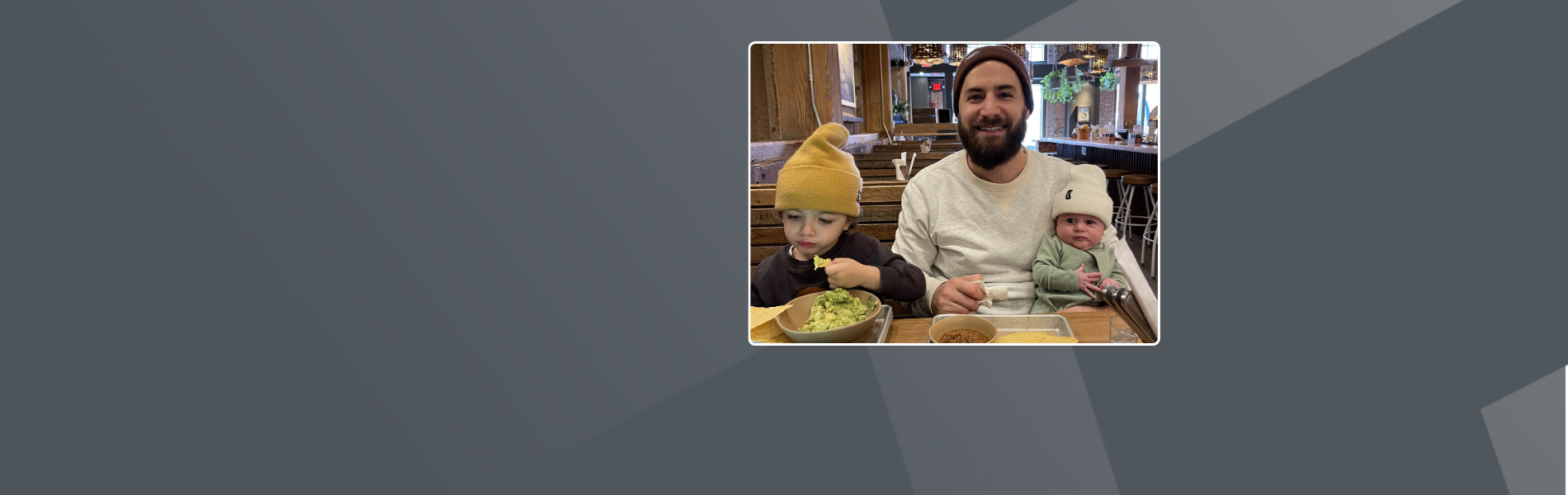 Steve, a white American man with his two children in a restaurant. He wears a hat and a white shirt, and has a beard. One child is on his lap and the other sits next to him eating.