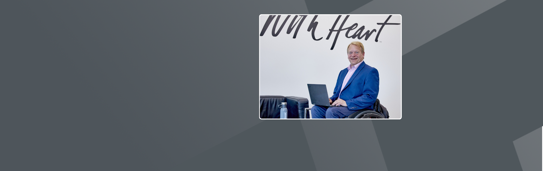 David, a white male Scott, sitting in an office in a black and red wheelchair with blonde hair, blue jacket blue and trousers with pink shirt, resting a laptop on his legs with a logo on the wall behind that says ‘with Heart’.