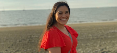 Stella, a woman in a red dress is standing by the sea.