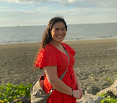 Stella, a woman in a red dress is standing by the sea.