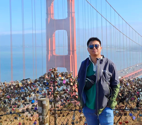 一位戴着墨镜的男士穿着一件外套，站在红色的大桥前