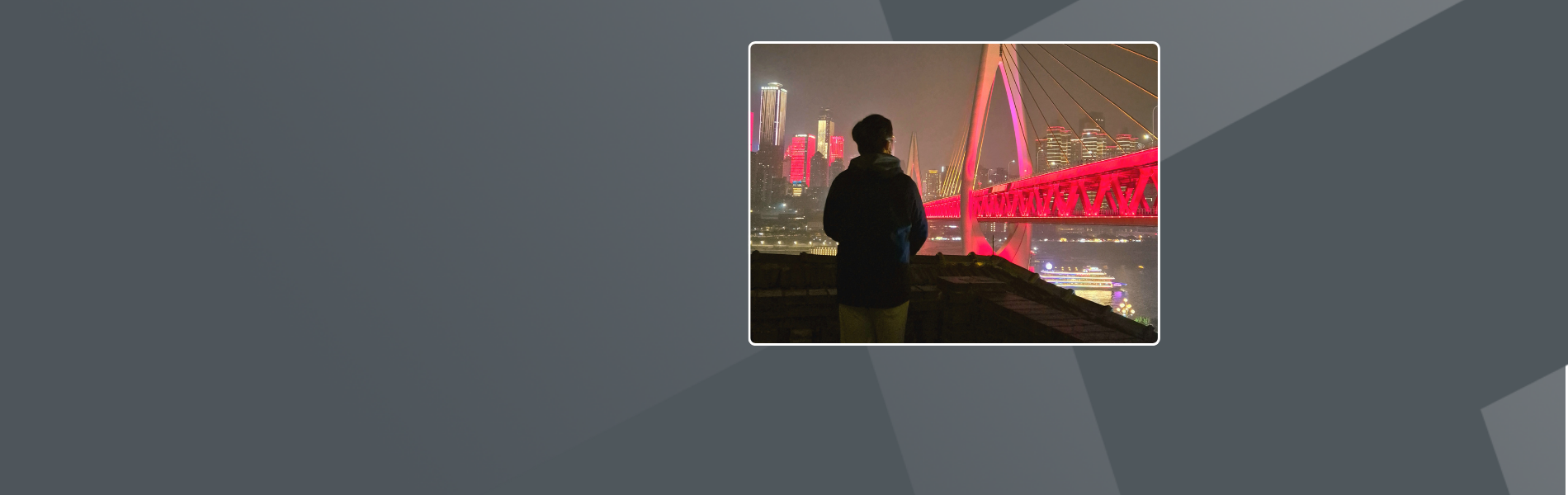 Brian, a short-haired wearing a jacket is looking at a bridge illuminated in red by lights and the distant night view. 