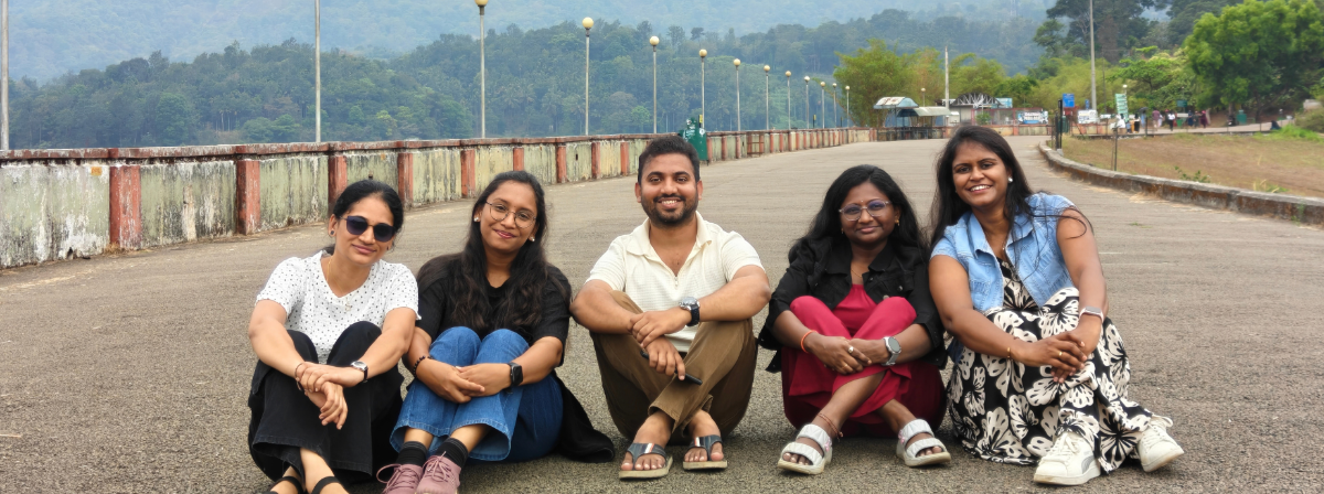 Five colleagues seated together outdoors on a wide road, with trees, hills, and a long walkway stretching into the background.