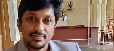 Ashok, Indian male, with dark hair and a trimmed dark beard and moustache. He wears a grey suit and grey shirt.