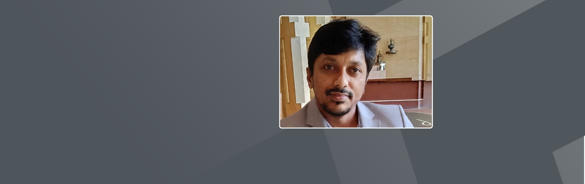 Ashok, and indian male, with dark hair, and a dark beard. He is wearing a grey suit with a grey shirt.