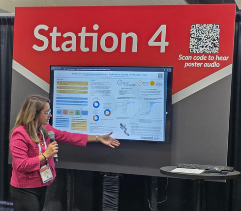 Diana, explaining a Parkinson's disease research poster. Digital display shows recruitment and retention trends in trials . Conference booth setting.