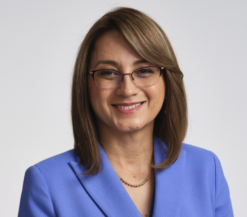 Diana, a Colombian woman, with shoulder-length brown hair wearing glasses and a vibrant blue blazer, smiling warmly against a light gray background.