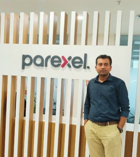 Hitarth standing indoors beside a wooden slat partition with the “parexel” logo mounted on a white panel. He is wearing a dark shirt and light-colored pants, with one hand in a pocket.