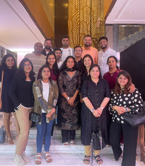 A group of people posing together indoors in a well-lit lobby area with marble flooring. The group is arranged in multiple rows, standing on steps near a decorative wall panel with gold patterns and a staircase visible in the background.