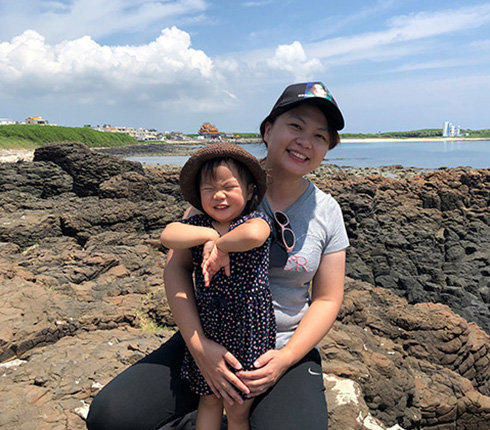 Joy Lin, Senior Director of Statistical Programming, is outside with her young child by the seaside. They are wearing outdoor attire with a cap and they are looking and smiling to the camera