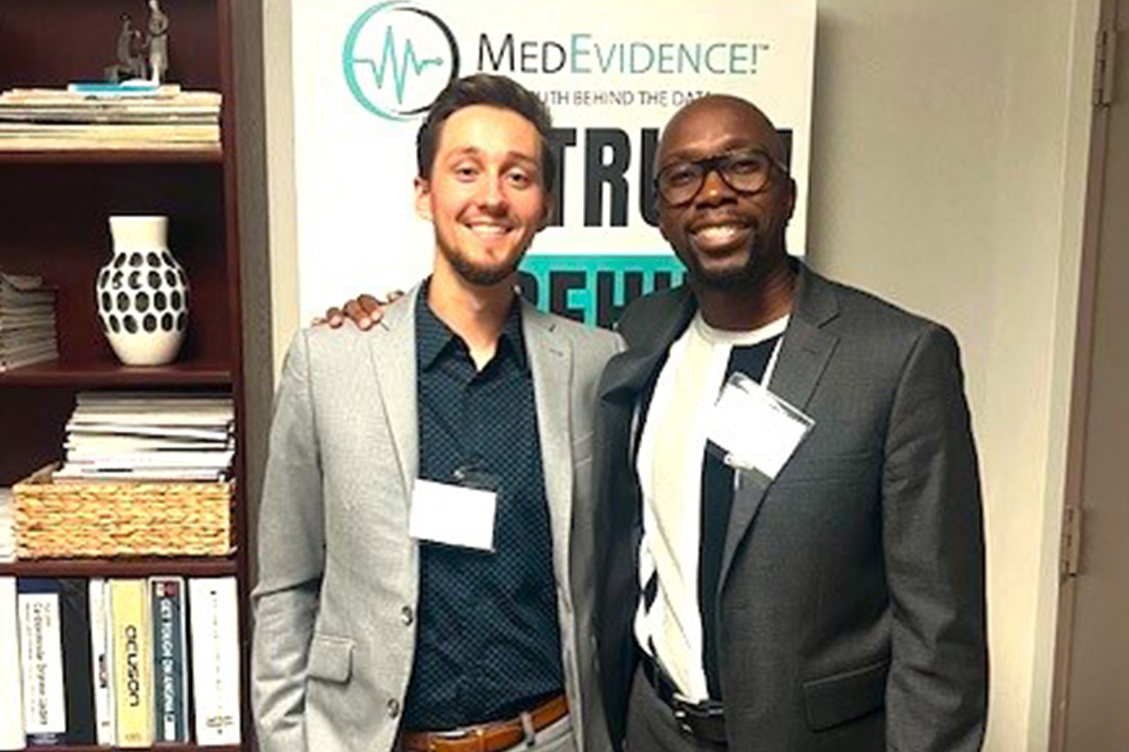 Two professionals working in the clinical operations industry is within an office, smiling at the camera. They are wearing business casual.