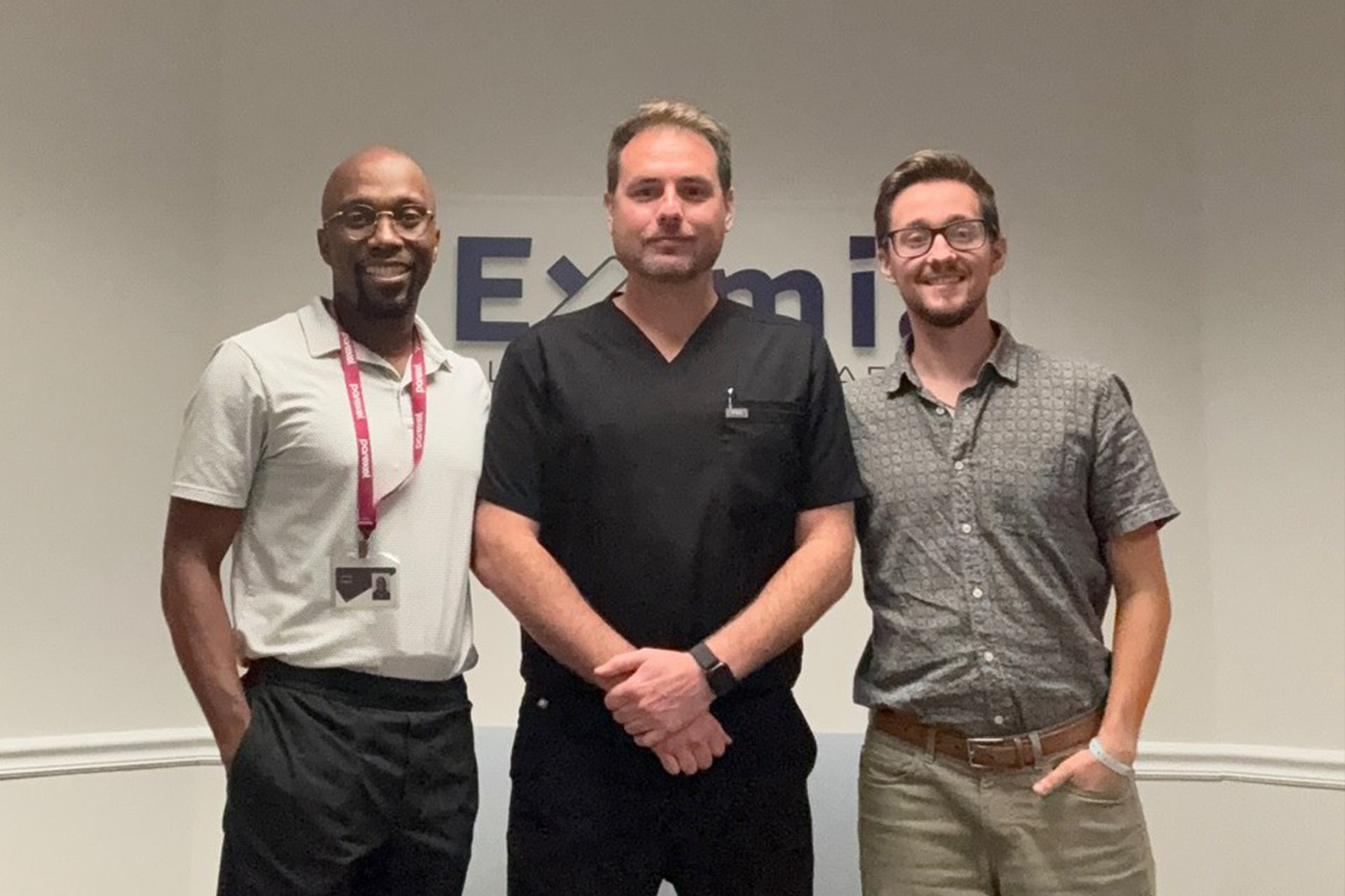 Three men working in the clinical operations industry are side by side wearing business casual. They are indoors and are smiling and looking into the camera