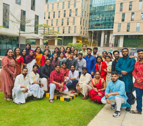 Large group of people posing outdoors on a lawn, dressed in colorful traditional attire with office buildings in the background.