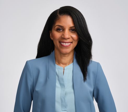 Professional headshot of Mika. She is smiling straight to the camera with a warm and confident expression. She has long dark hair, wears a light blue blazer and matching blouse against a neutral gray background.
