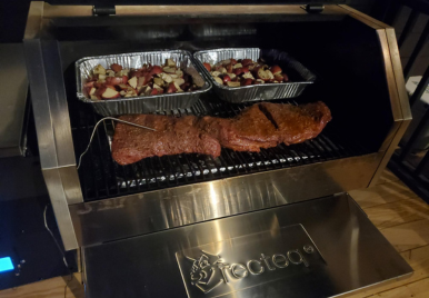 Stainless steel pellet grill with open lid displaying three large ribeye steaks on the main grate and two aluminum trays of vegetables on the upper warming rack, set on a wooden deck.