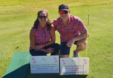 Ryan and wife kneeling on golf course in matching pink leopard print outfits with memorial signs honoring family members at cancer awareness charity event.