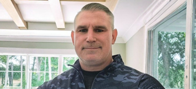 Headshot of Ryan, a smiling Caucasian man with short silver-gray hair and trimmed goatee wearing dark camo jacket.
