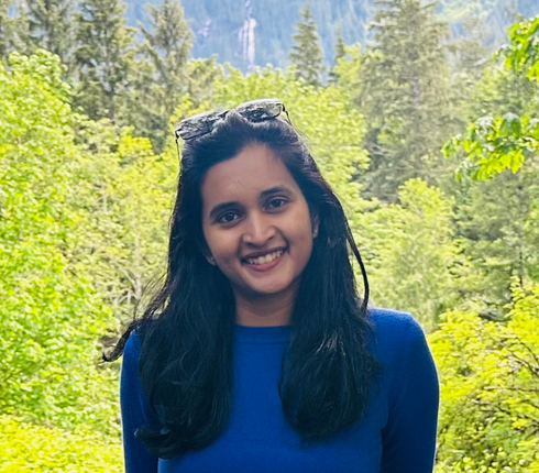 A Woman, Sathvika, standing outdoors in front of dense green trees and tall mountains in the background, wearing a blue long‑sleeve top with sunglasses resting on the head.