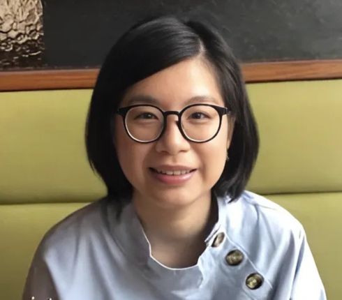 Tina, a Taiwanese woman, with short dark hair wearing round glasses and a light-colored top with decorative buttons, sitting against a green upholstered booth.