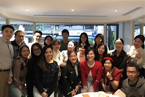 Parexel Taipei team picture. The team is gathered in a bright office space with large windows. People are arranged in two rows, wearing business casual attire in various colors, smiling at the camera.