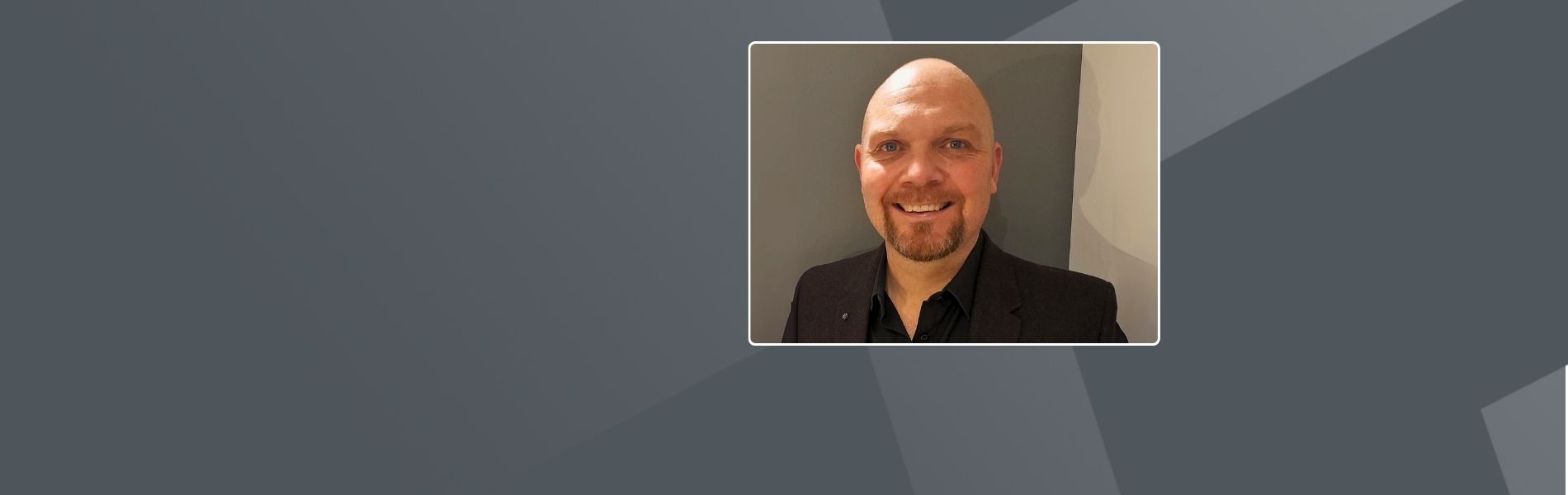 Tom, and English man, with a bald head and ginger beard, smiling warmly at the camera. He is wearing a black suit jacket and black button-up shirt against a gray background.