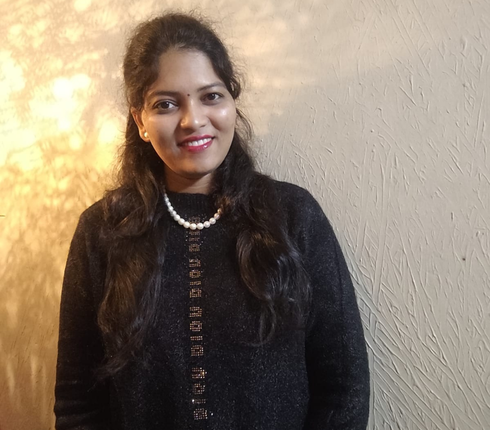 A woman standing indoors against a textured beige wall, smiling at the camera. She has long dark hair and is wearing a black sweater and a pearl necklace.