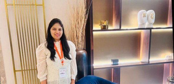 A woman standing indoors near decorative shelving with soft lighting. She is smiling, wearing a light-colored patterned blouse and an orange lanyard.