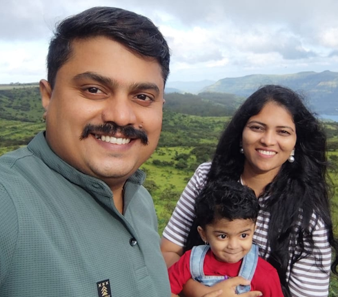 A group of three people outdoors in a lush green hilly landscape. One person is holding the camera and standing in the foreground. Another person is standing beside them, and a small child is positioned between the two adults.