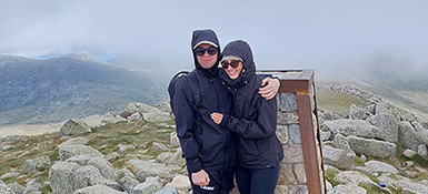 Adrian Cawley, an Australian male, Site Care Partner (SCP) and a woman are captured on top of a majestic mountain, both clad in durable navy outdoor gear and sporting sunglasses.
