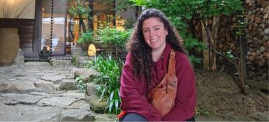 Andrea, a Spanish woman, with long curly hair smiles brightly. She sits in a Japanese courtyard.