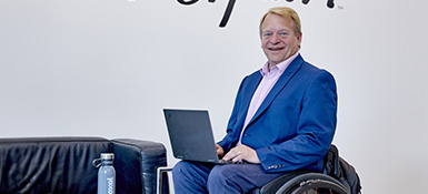 David, a white male Scott, using a wheelchair sits with a laptop on their lap. He is wearing a light pink shirt, blue jacket, and jeans. The Parexel tagline 'With Heart' appears in the background.