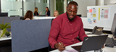 Henry, a Black American Clinical Research Associate, is focused on his work at a desk, utilizing a laptop. He has short hair, a beard, and a red shirt.