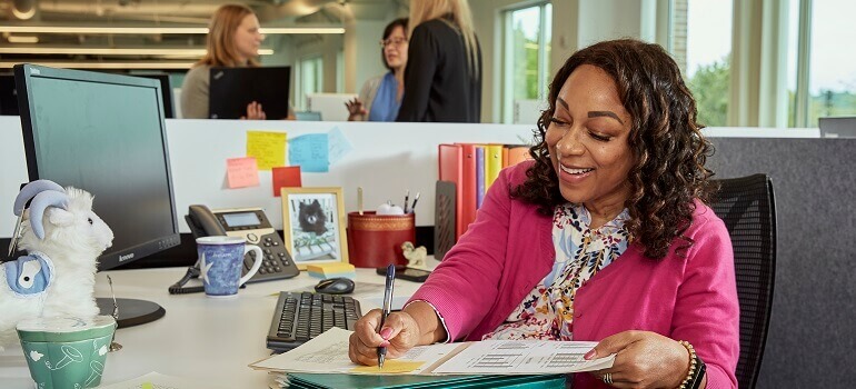 Sheila, a Black American Clinical Operations Leader at Parexel, sits at a desk, writing in a clinical trial document. She has shoulder-length curly hair and is wearing a pink cardigan and patterned shirt.