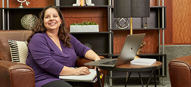 Cristina, a female Italian-American, is a Clinical Operations Manager at Parexel. She is seen working from home, with a laptop on a table and notebook on her lap.