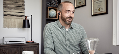Middle-age white Male working from home. He has a headphone in his ear, looks at the screen and is smiling. He wears a plaid shirt, has a beard and short hair.