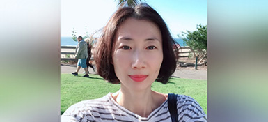 Inhye, a female Korean, with chin long dark hair, red lipstick and a striped shirt taking a selfie with a beach walk in the background.