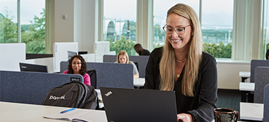 Jessi, a white American Finance professional, is working at a common table in an office. Engaged in her tasks, she writes in a notebook with a cup placed nearby. She has long blond hair and wears a dark shirt.